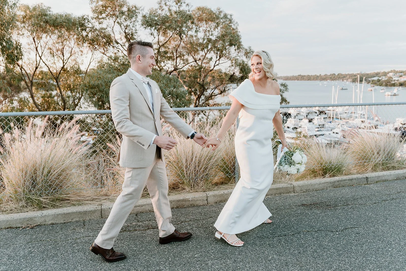A bride and groom holding hands and smiling, walking near a waterfront with trees and boats in the background.