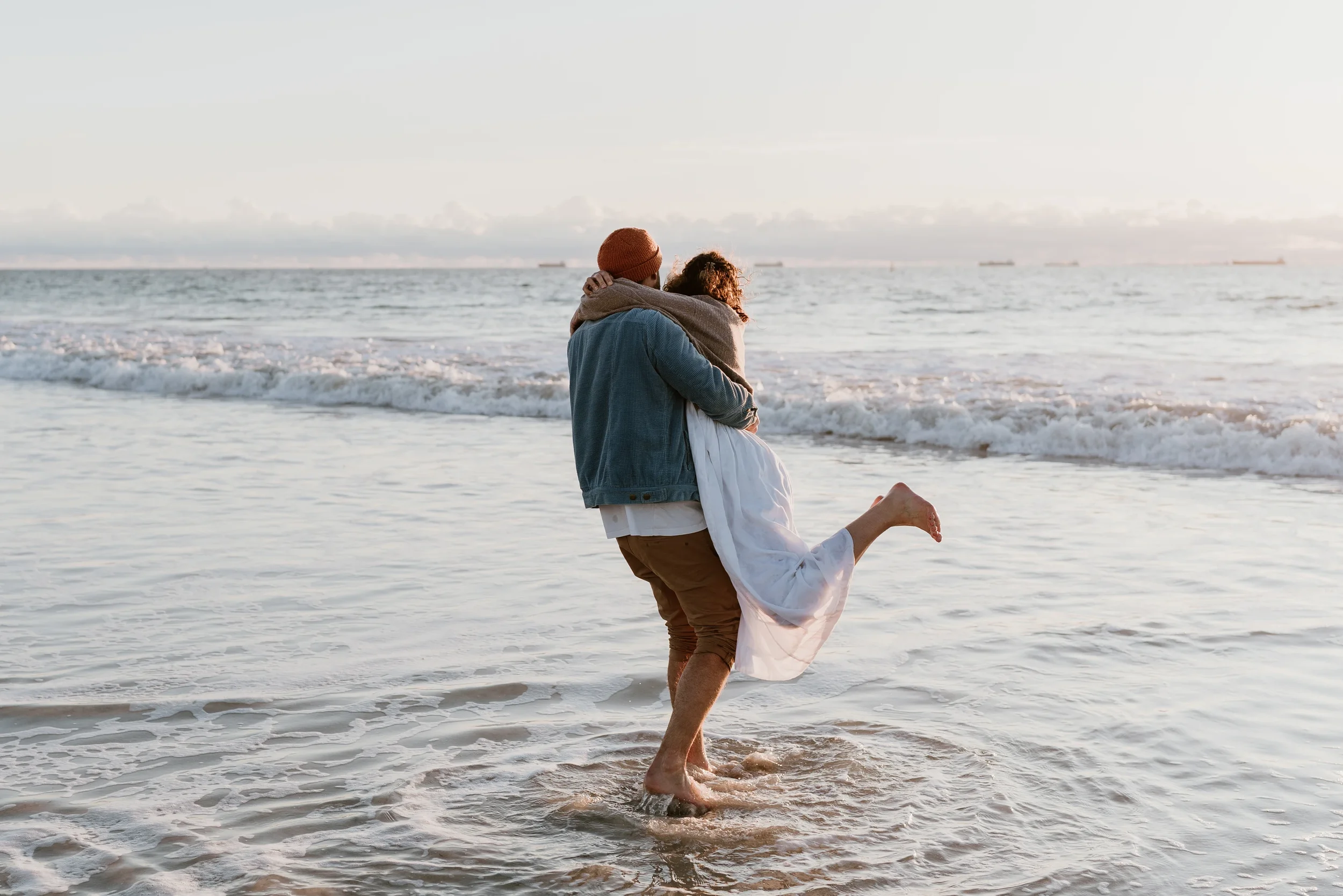 A couple hugging on the beach with the man carrying the woman, both standing in shallow water, during sunset.