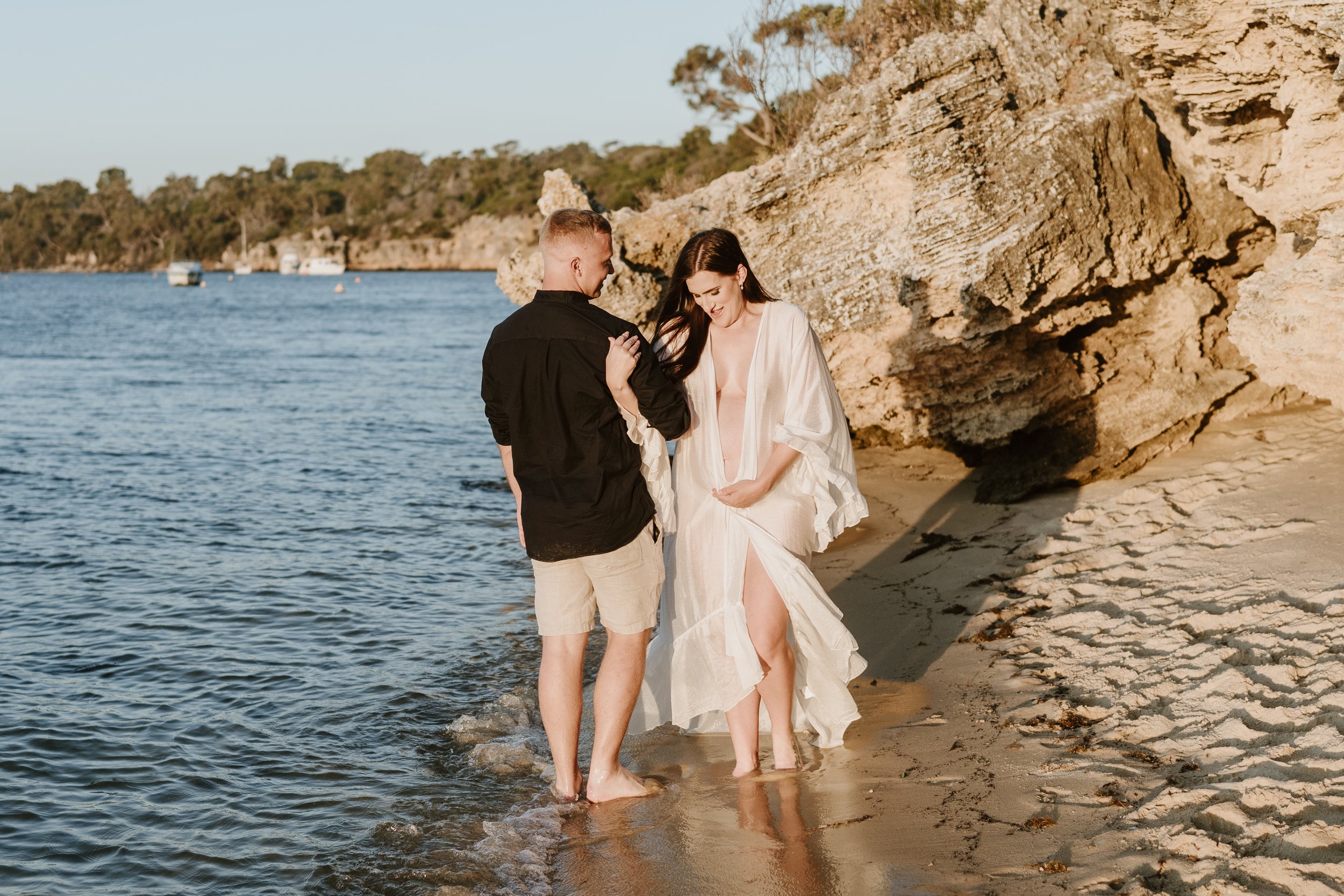 A couple enjoying a romantic moment at the beach during sunset, with the woman in a flowing dress and the man in a black shirt and shorts, standing in shallow water near rocks.
