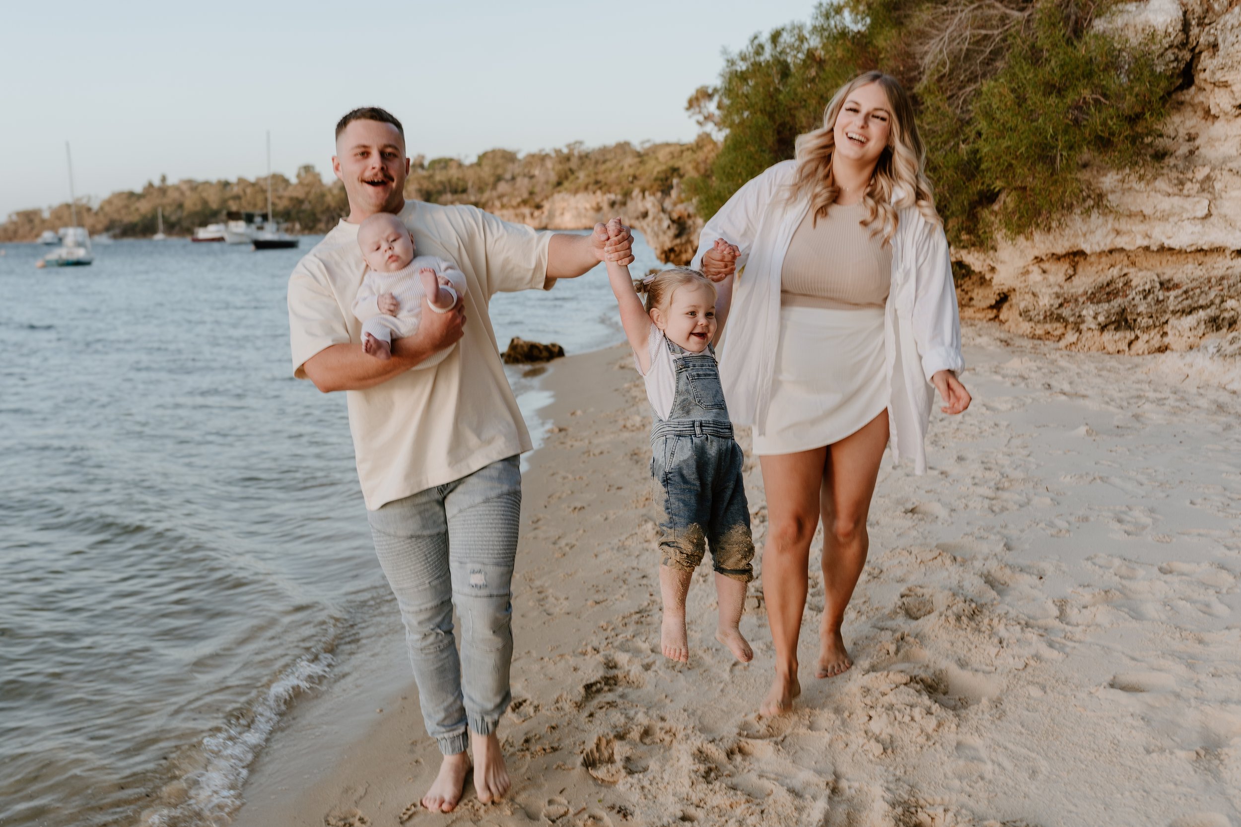 A family of four walking on a sandy beach, holding hands, with sailboats in the water and rocky cliffs with greenery in the background.