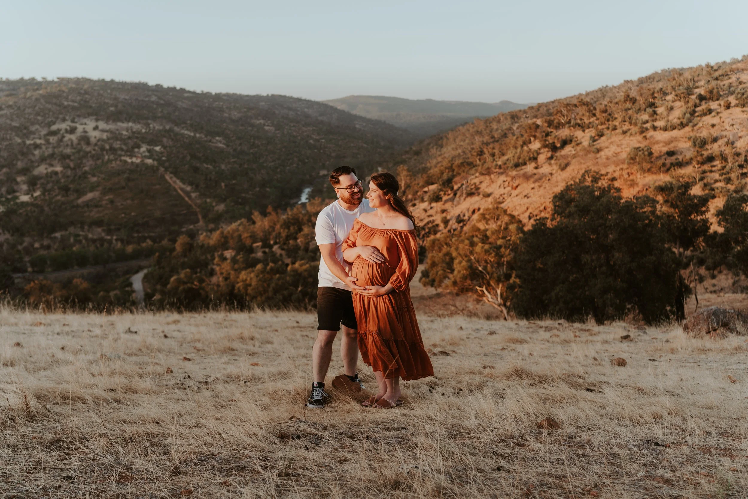 A pregnant woman and her partner standing in a field at sunset, smiling and holding her belly against a scenic mountainous landscape.