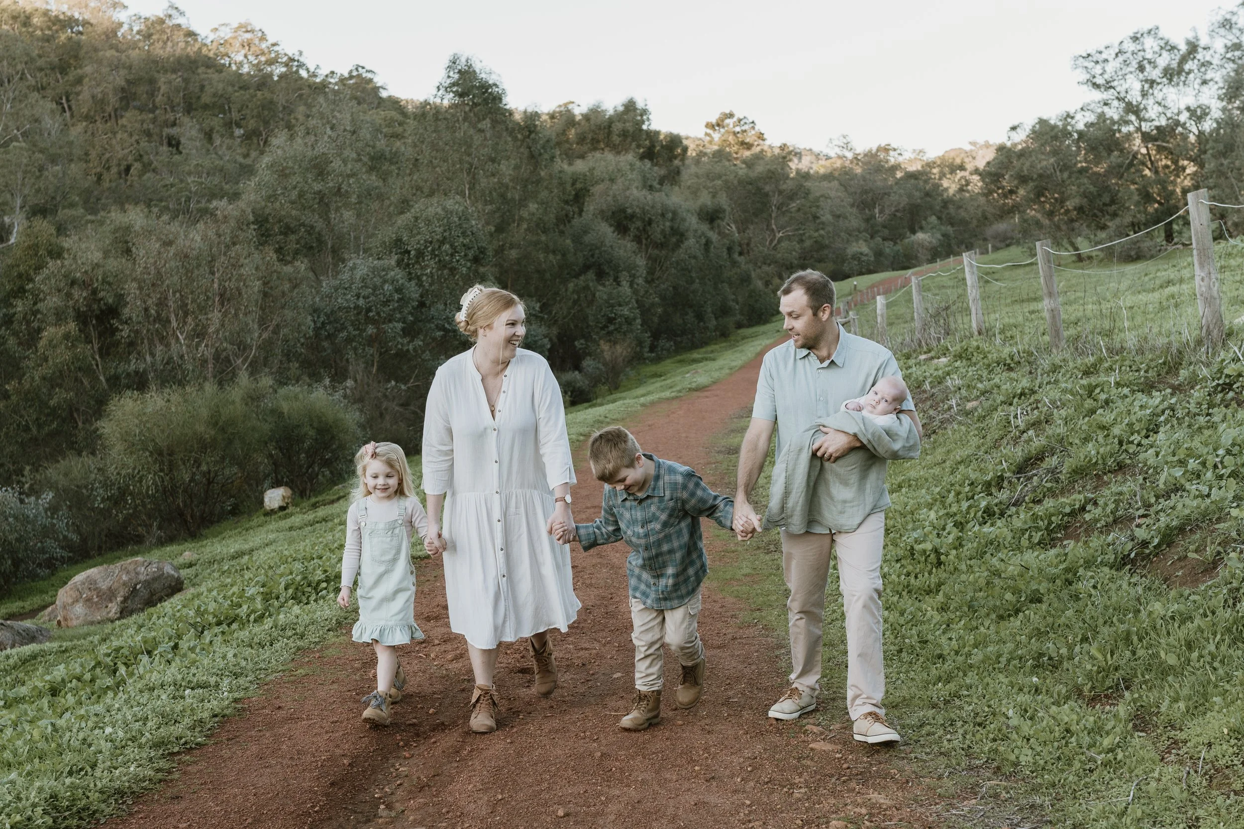 A family of six walking on a dirt trail in a grassy, wooded area during daylight. The family includes two adults, three children, and a baby, all smiling and holding hands.