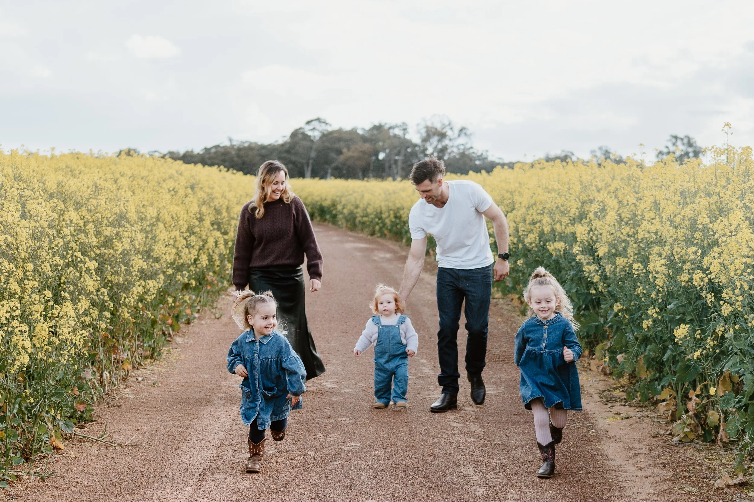 Family of five walking and playing on a dirt path surrounded by yellow flowering plants, with a cloudy sky above.