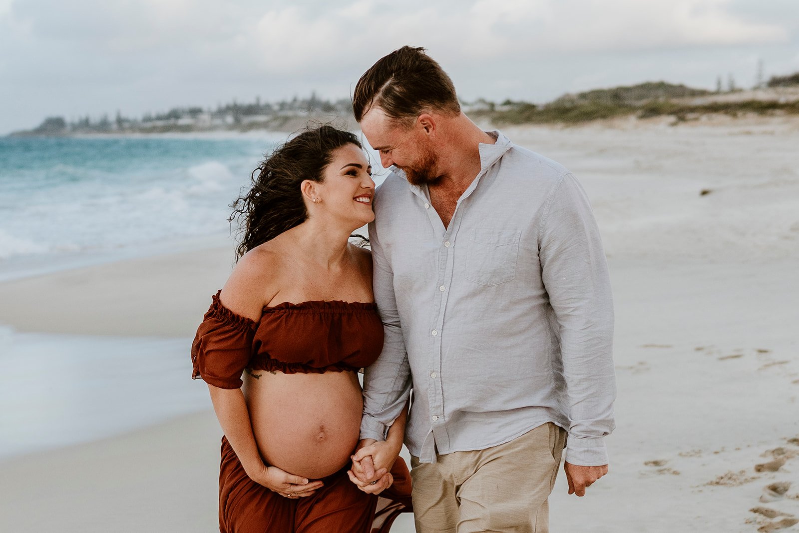 A pregnant woman and a man holding hands on the beach, smiling and looking at each other.