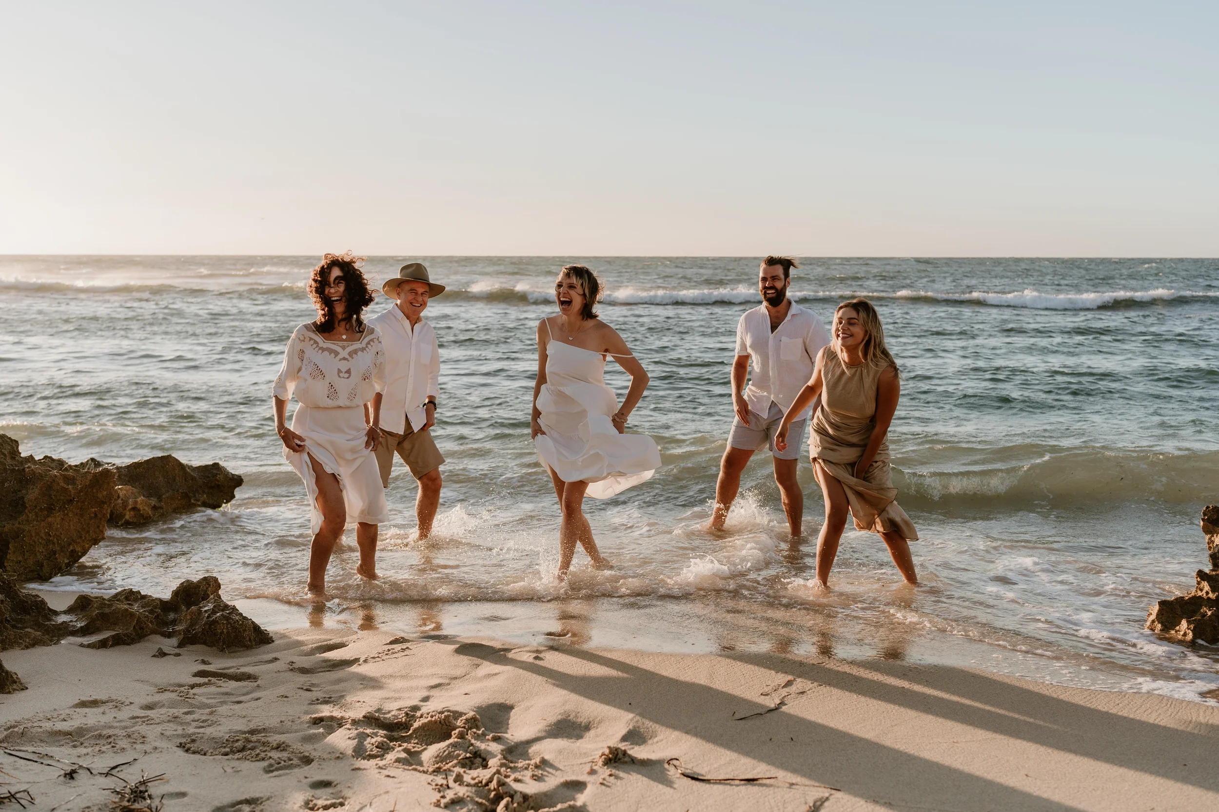 A family laughing together in the shallows at the beach during their photoshoot session, in coordinated neutral coloured outfits that complement the sand and sea