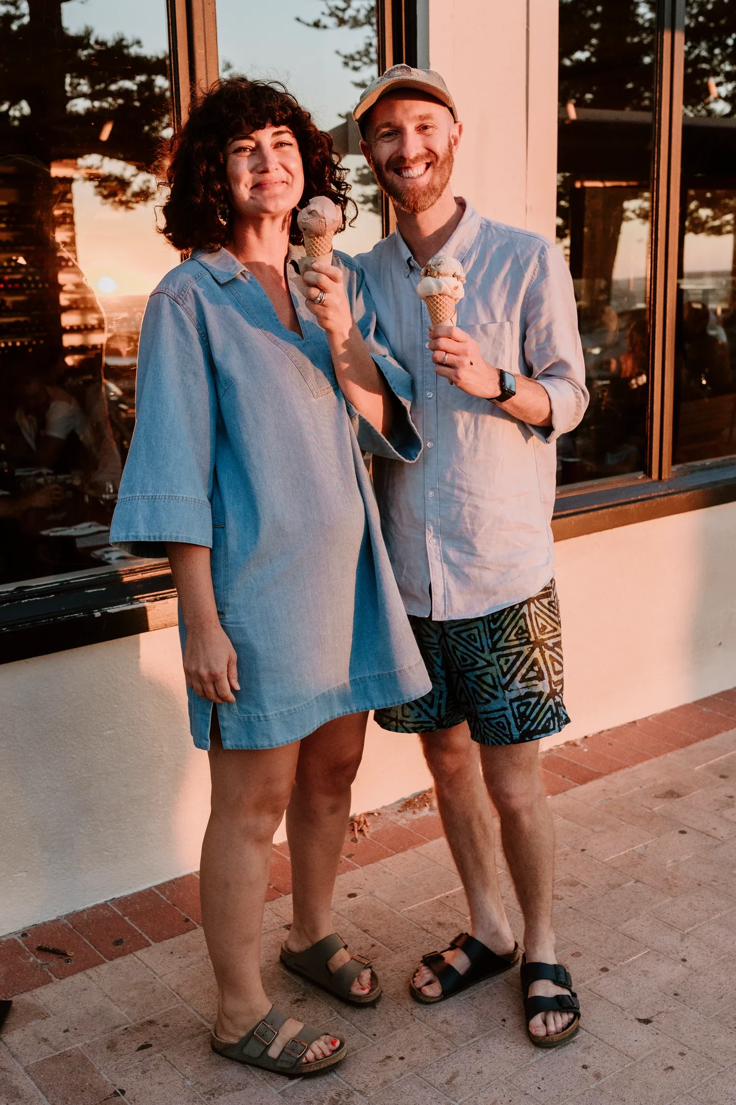 A woman and a man smiling and holding ice cream cones outdoors during sunset, standing on a brick patio near a building with large windows.