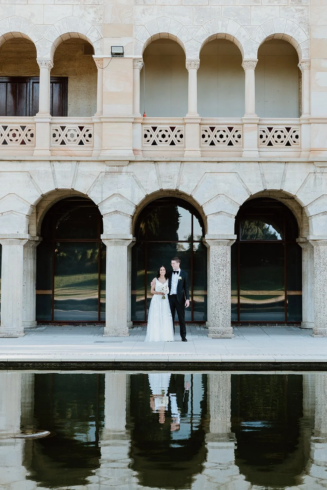 A bride and groom in formal wedding attire standing together in front of large glass doors with reflections of greenery, at a building with arched architecture and balcony, reflected in a water feature in the foreground.