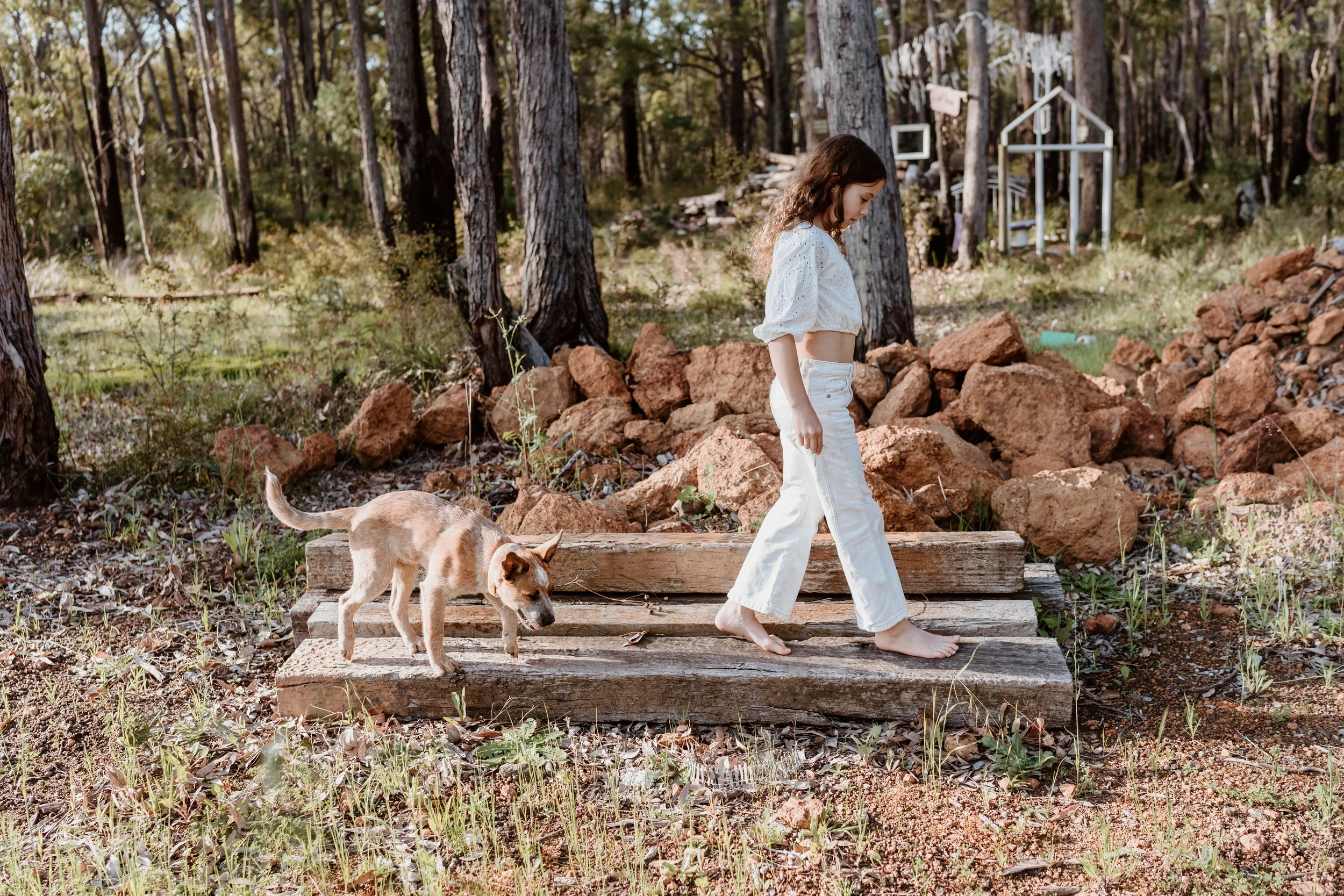 A young woman with curly hair wearing a white crop top and white pants walking barefoot on a wooden plank bridge in a forested area, followed by a small dog with a light brown and white coat. In the background are trees, rocks, and a small white meta