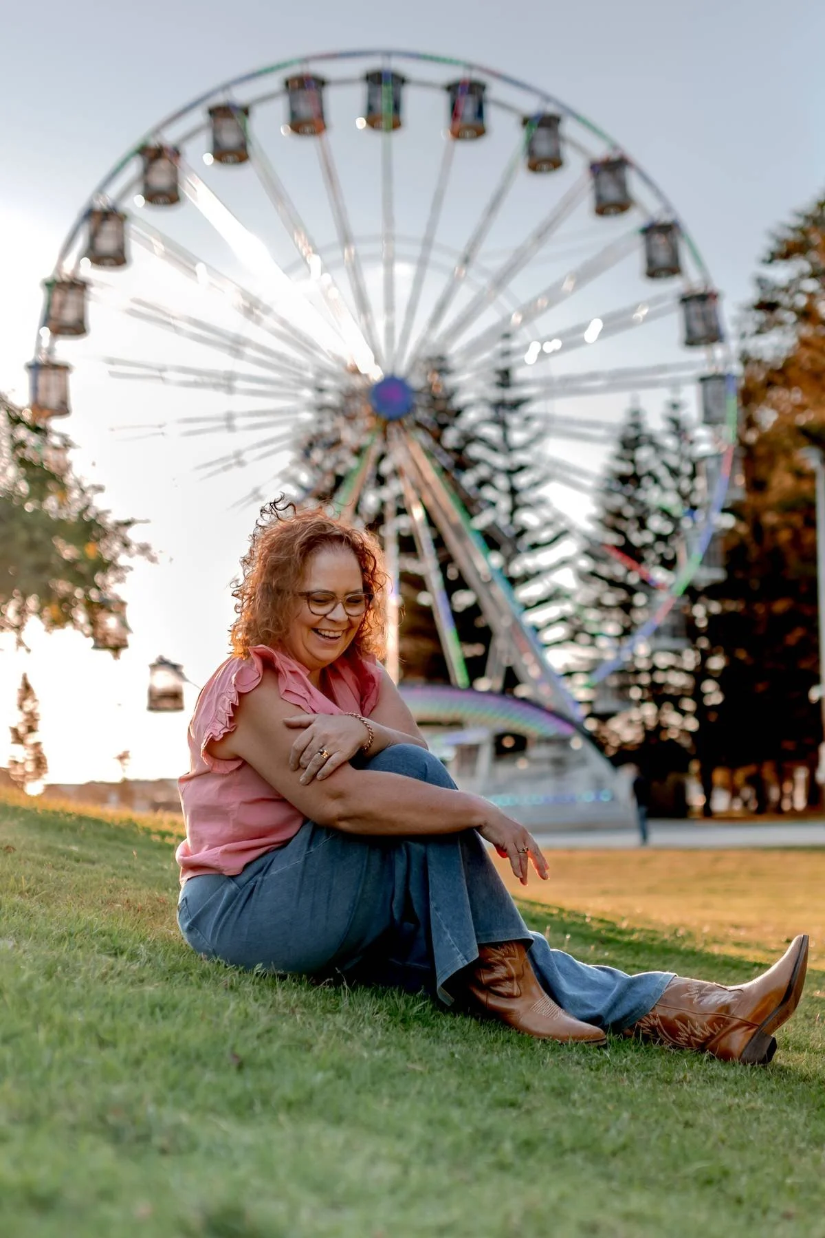 A woman sitting on grass in front of a large Ferris wheel, smiling and hugging her knees, during the daytime.