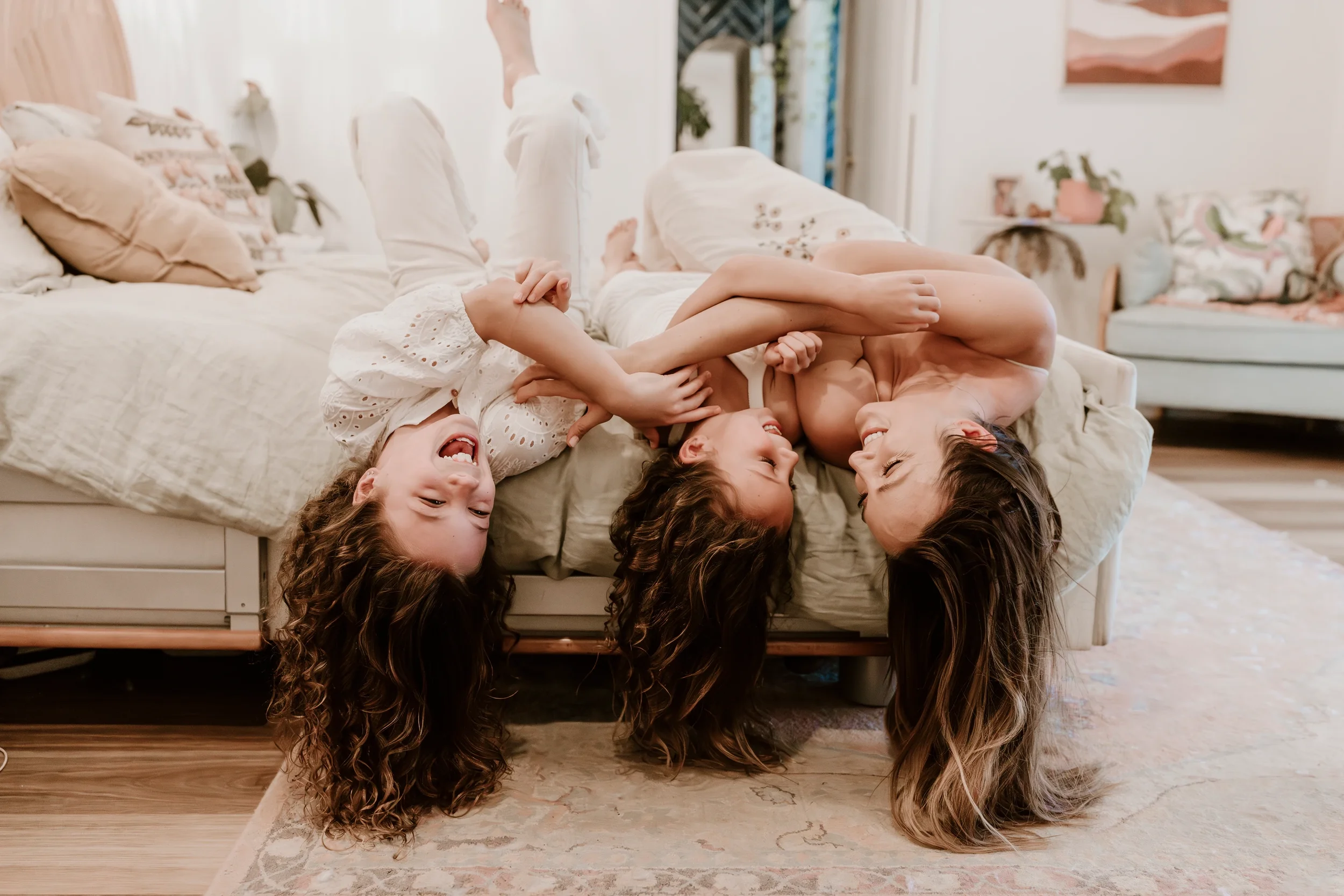 A woman and two children are playing and laughing together on a bed in a cozy living room.