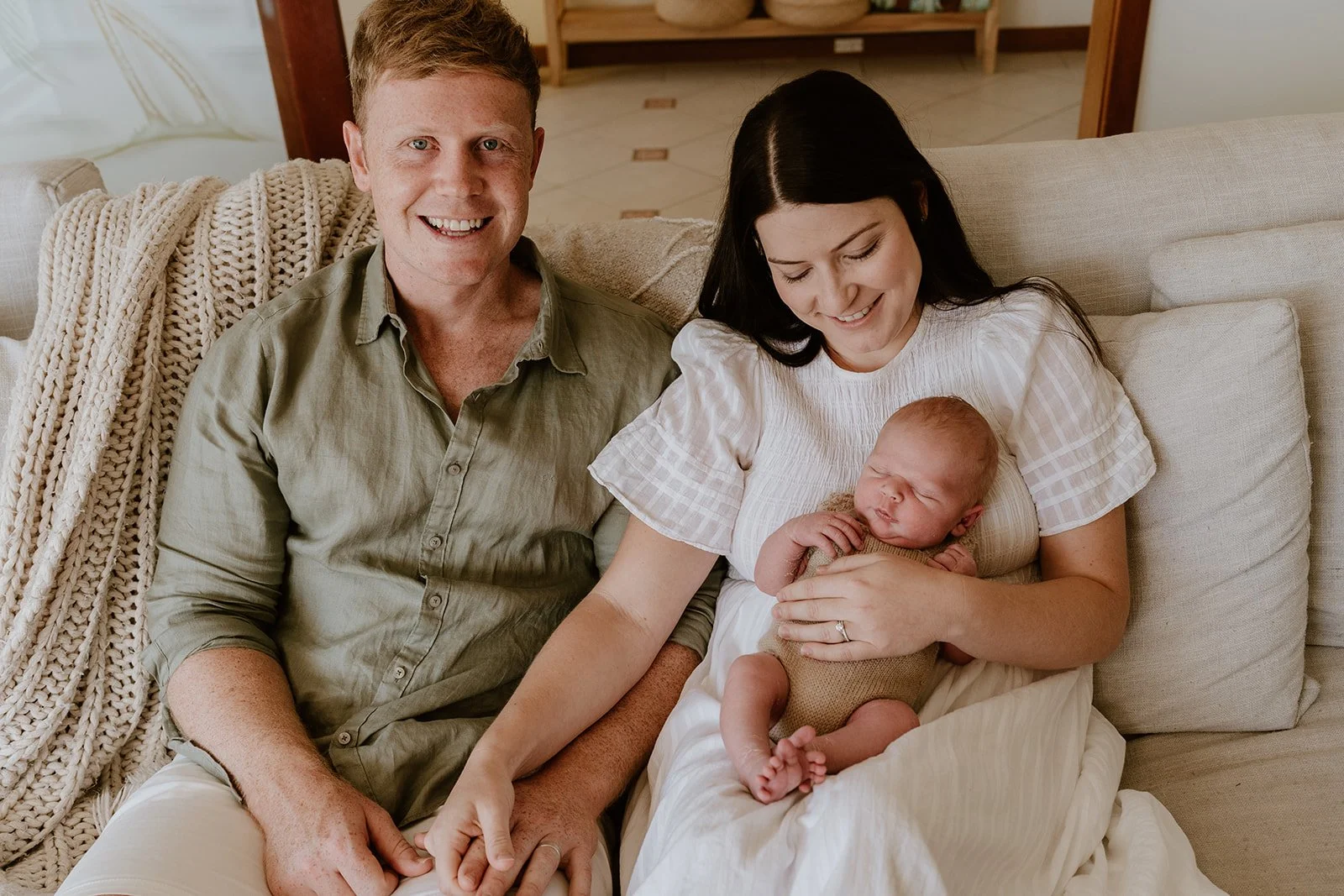 A family of three sitting on a beige couch; a woman holding a sleeping newborn, a man sitting next to her, smiling at the camera.