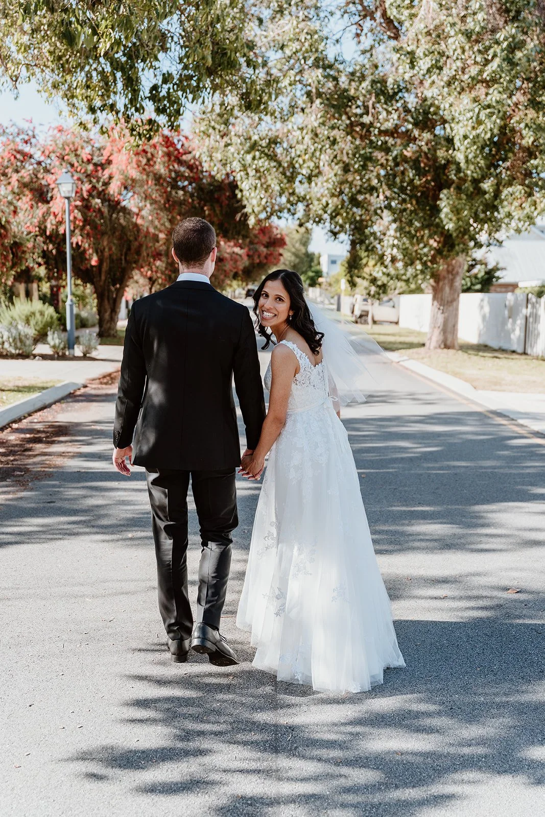A newlywed couple, the bride in a white lace wedding dress and veil, and the groom in a black suit, walk hand-in-hand down a quiet street lined with trees and greenery, with the bride smiling at the camera.
