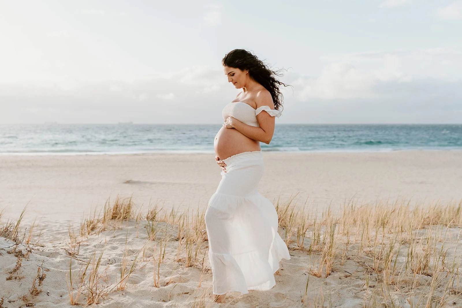 Pregnant woman standing on a sandy beach near the ocean, wearing a white off-shoulder top and long white skirt, holding her belly and looking down, with a cloudy sky in the background.