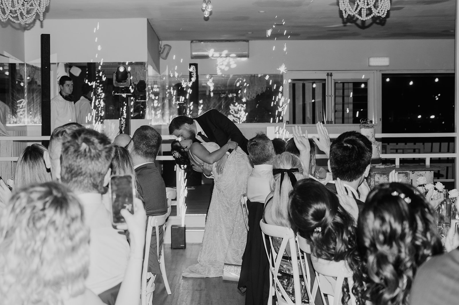 Couple dancing and kissing at a wedding reception with guests clapping and cheering, sparklers in the background, in a decorated indoor venue.