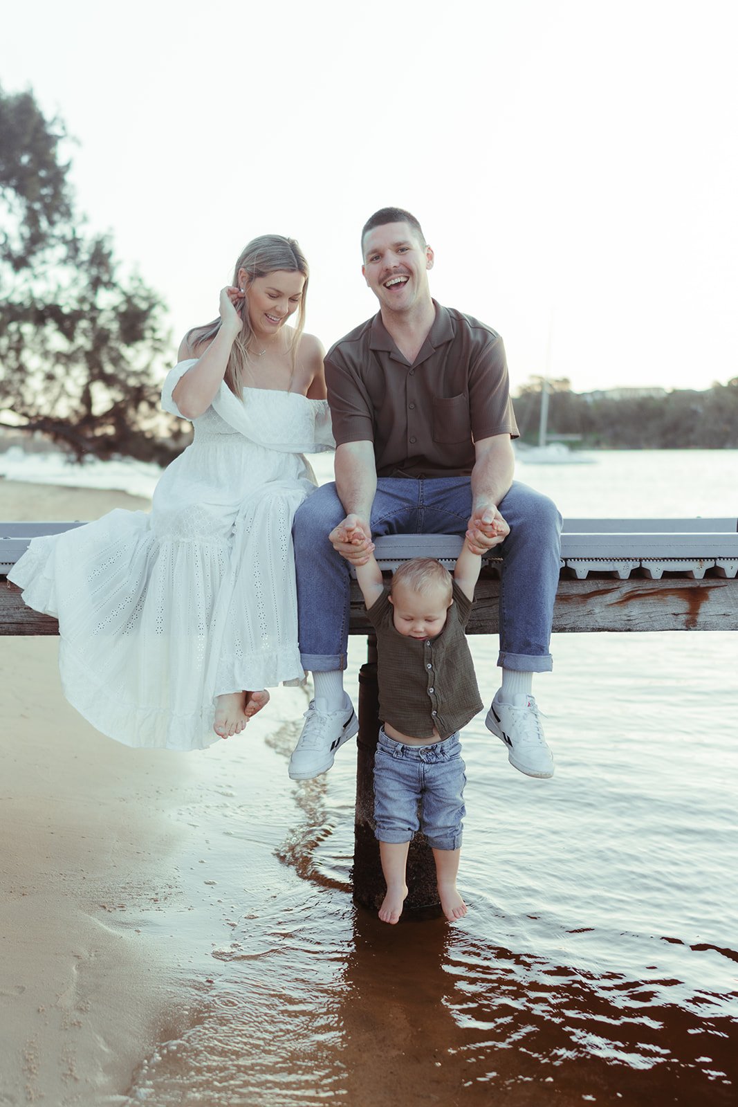 A family of four enjoying time at the beach near water. The mother is wearing a white dress, seated on a wooden dock, smiling, with her hand touching her hair. The father, dressed in a dark shirt and jeans, is sitting next to her, holding their young