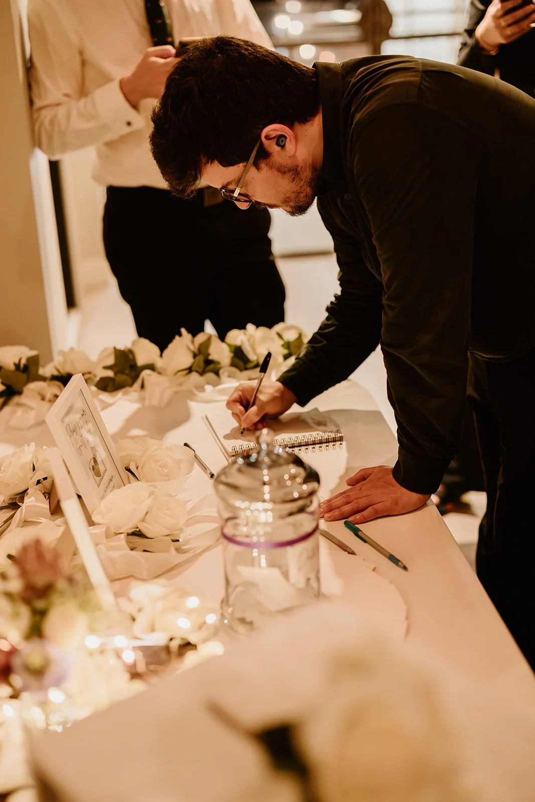 A man in black clothing signing a guestbook at a table decorated with white roses and framed photos, surrounded by other people.