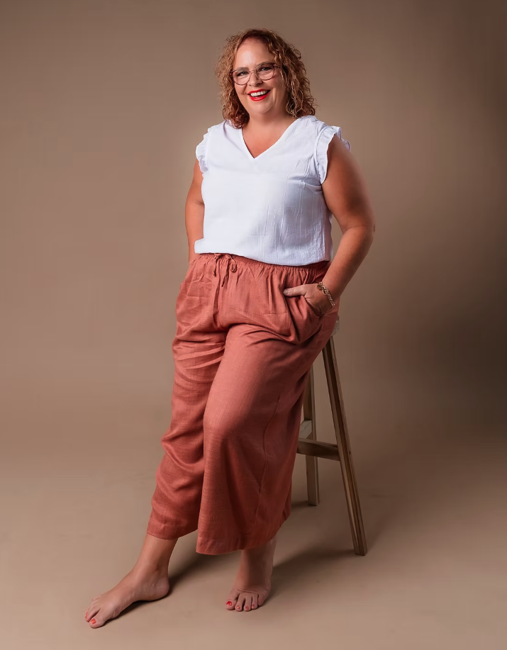 A woman with curly red hair and glasses, wearing a white sleeveless top and loose pink pants, standing barefoot next to a wooden stool on a beige background, smiling at the camera.