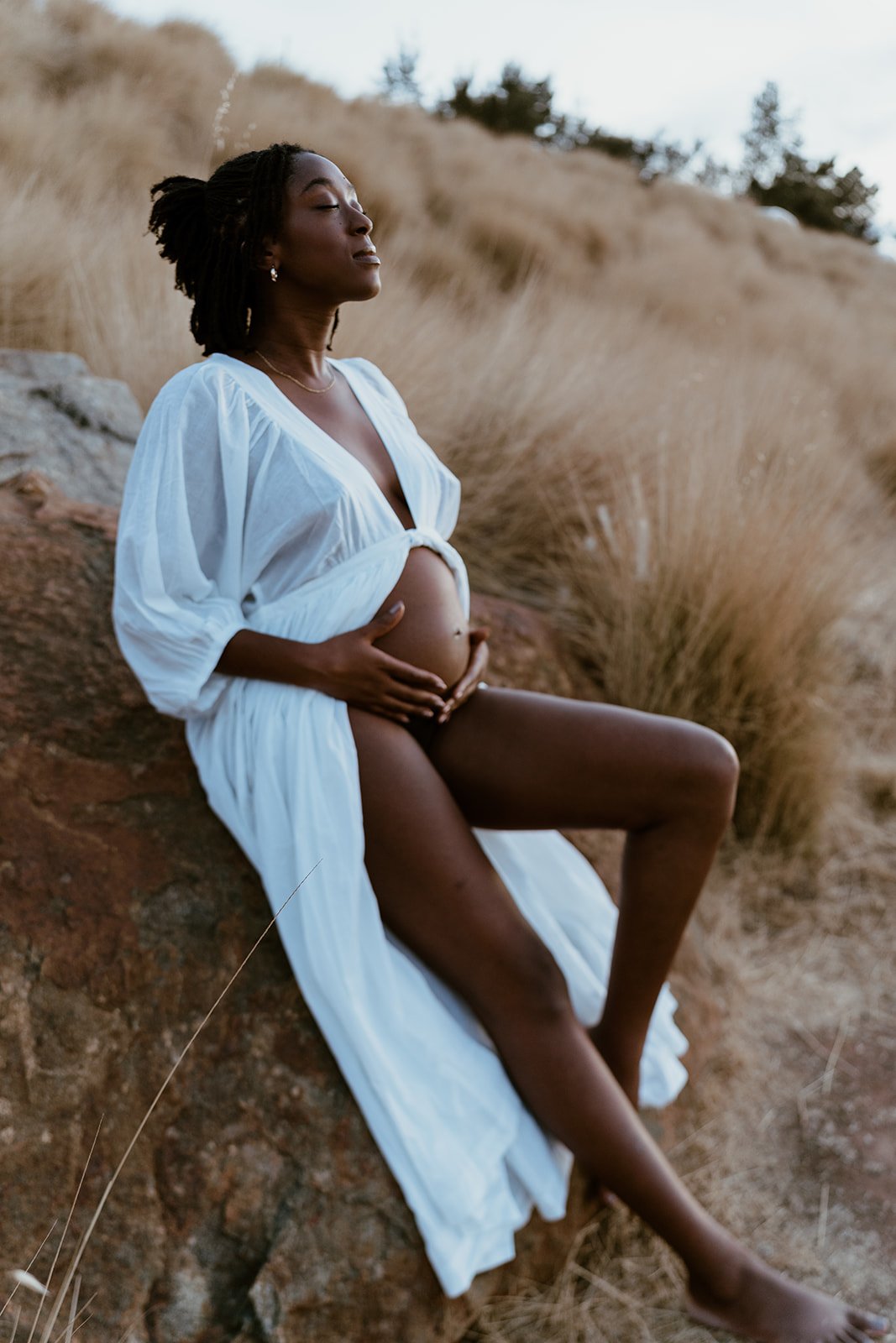 Pregnant woman sitting on a rock in a field with tall grasses, wearing a white dress, with closed eyes and a peaceful expression.
