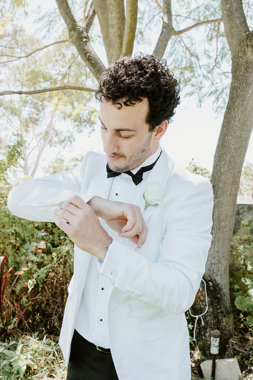 A man dressed in a white tuxedo with a black bow tie and a white boutonnière looks at his watch outdoors next to trees and plants.