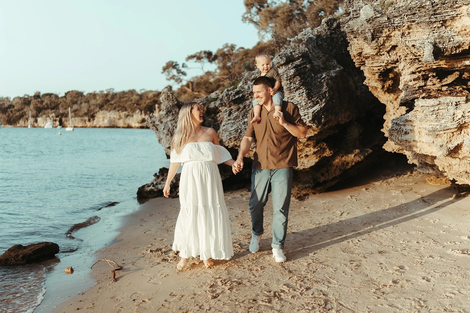 A family of three enjoying a walk on the beach during sunset, with rocky cliffs in the background. The man is carrying a young boy on his shoulders, and they are holding hands with a woman in a white dress. All are smiling and looking happy.