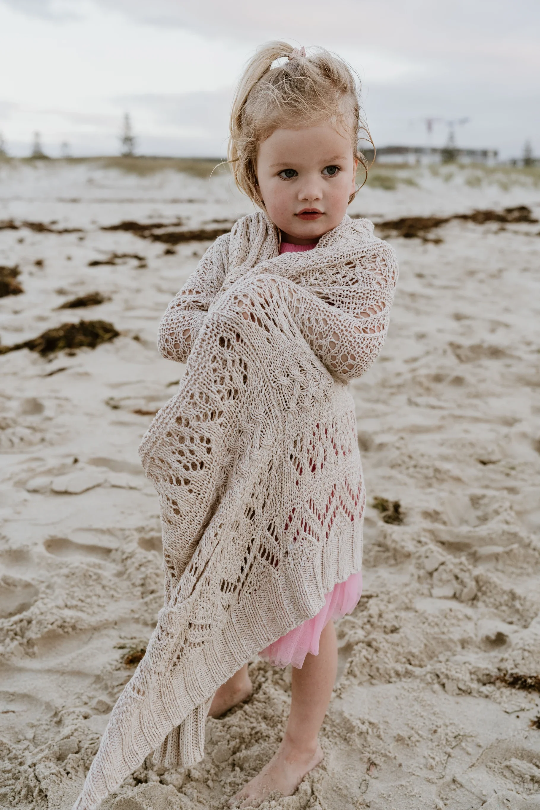 A young girl standing barefoot on a sandy beach, wrapped in a beige knitted blanket, with an overcast sky and power lines in the background.