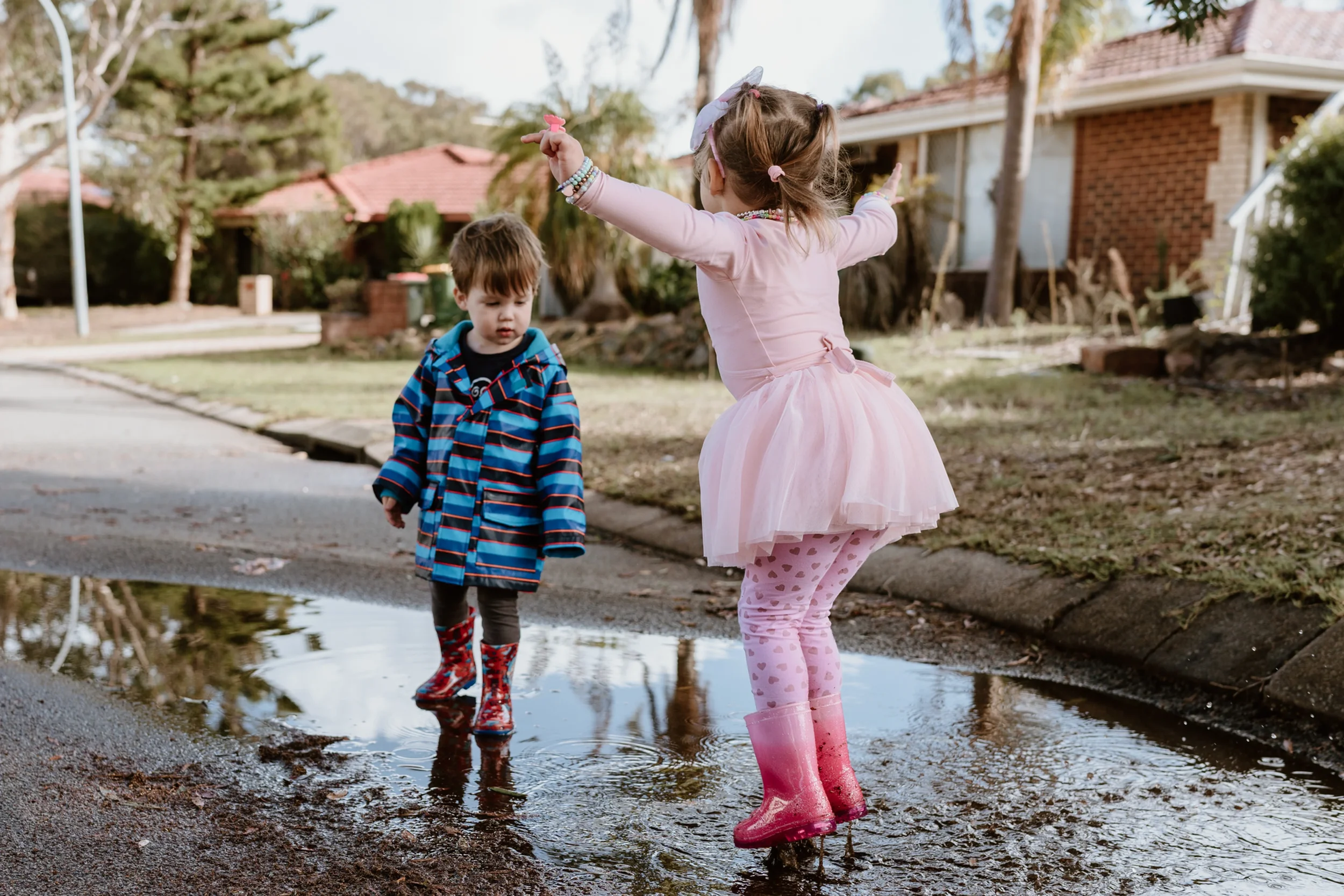 Two young children playing in a puddle on a residential street. The girl is wearing a pink dress, pink leggings with hearts, and pink rain boots, with her arms raised. The boy is wearing a blue striped jacket and red rain boots, looking at the puddle