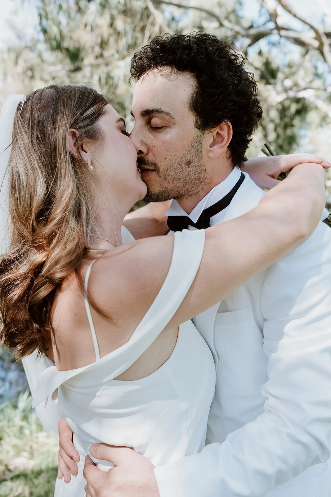 A bride and groom sharing a kiss outdoors, with the bride in a white dress and the groom in a white tuxedo jacket, surrounded by greenery.