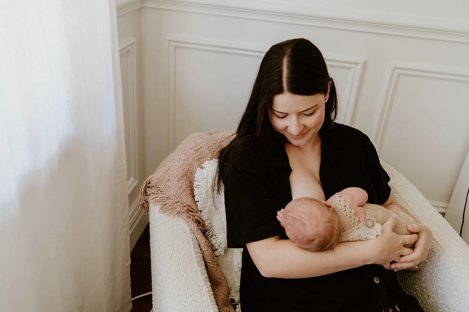 A woman with long dark hair breastfeeding a newborn baby in a cozy room.