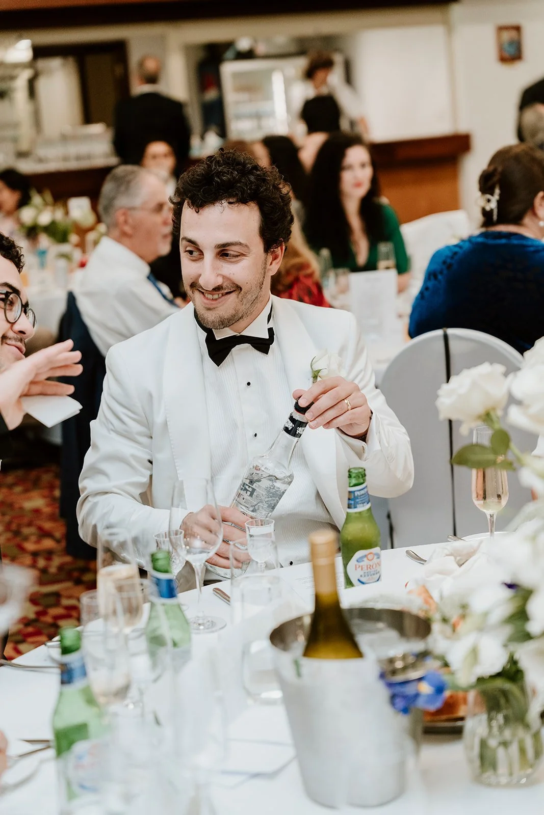 Man in white tuxedo pouring a drink at a wedding reception surrounded by guests and floral arrangements.
