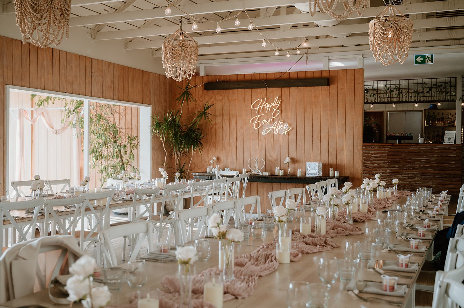 Wedding reception setup with long tables decorated with pink cloth runners, white flowers in glass vases, candles, and place settings. Wood-paneled walls, string lights, and a neon sign reading 'Happily Ever After' are in the background, with large windows and hanging chandeliers.