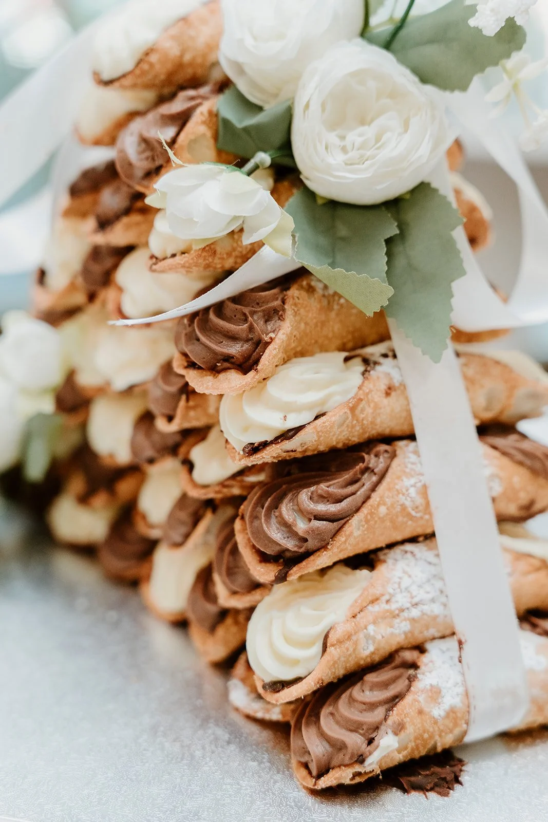 A stack of assorted cannoli, topped with white and cream-colored flowers and green leaves.
