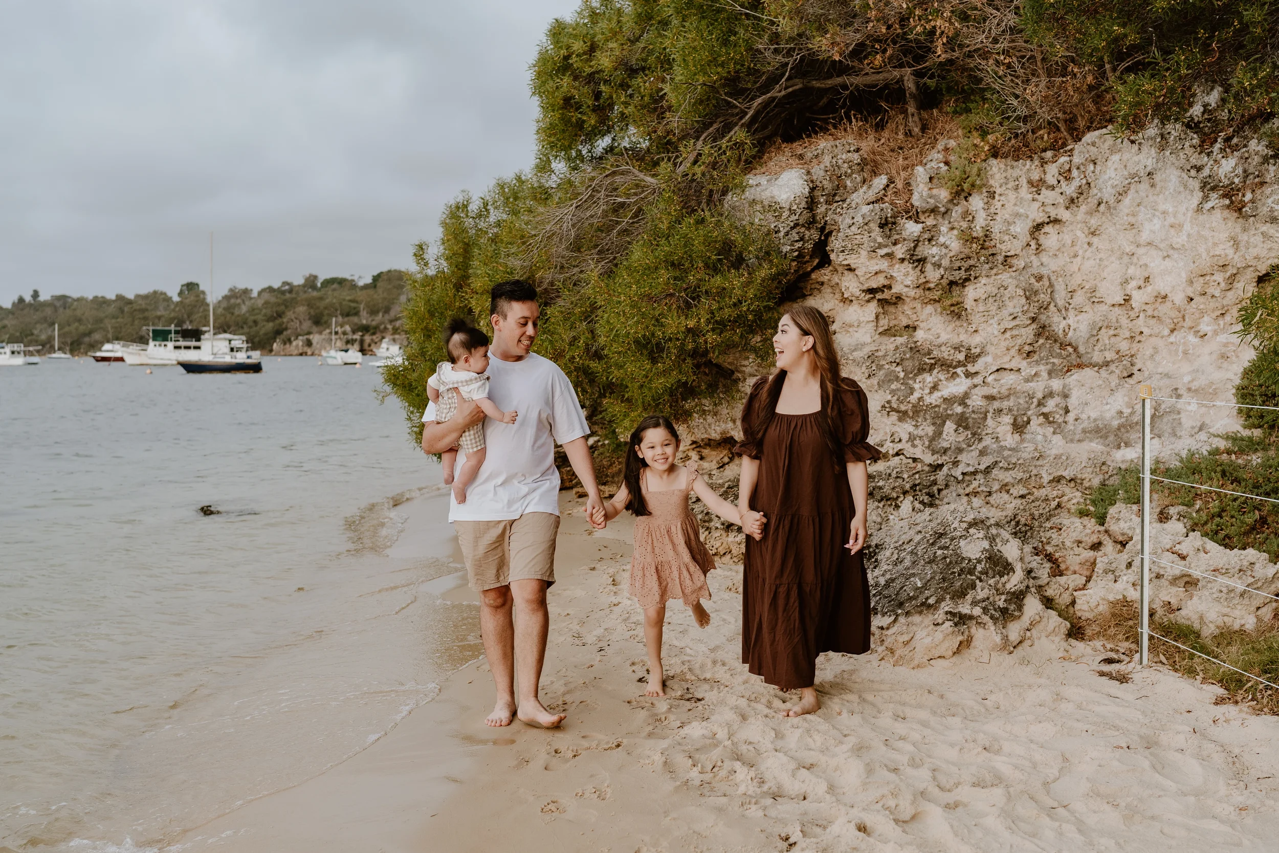 A family of four walking along a sandy beach holding hands, with boats anchored on the water and a rocky, tree-covered cliff in the background.