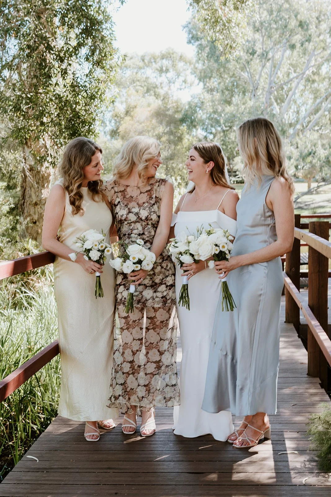 Four women, dressed in formal attire, standing on a wooden bridge outdoors, holding bouquets, smiling and enjoying a moment together.