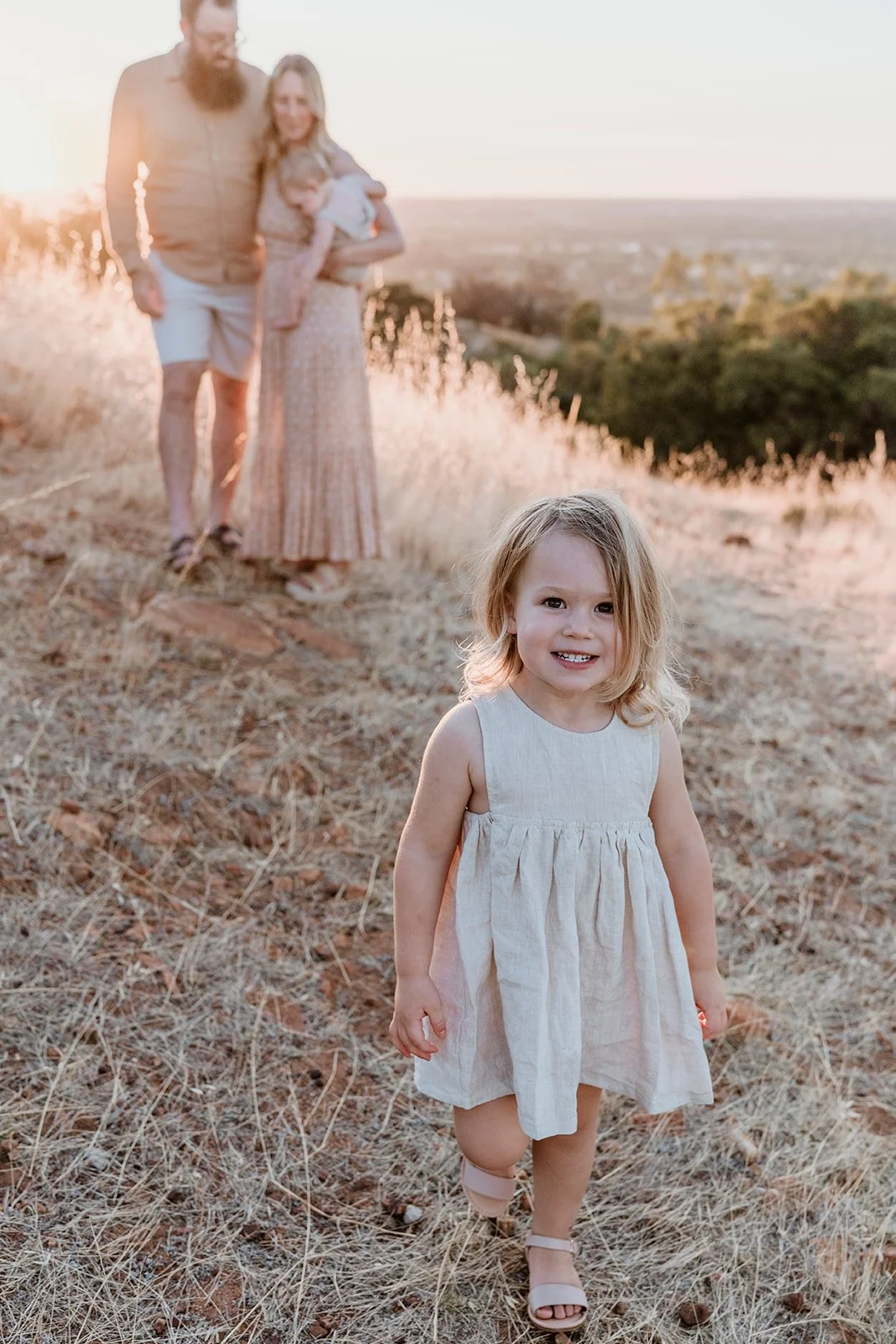A young girl with blonde hair smiling and walking on dry grass in a beige dress, with a blurred background of a couple and a sunset view.