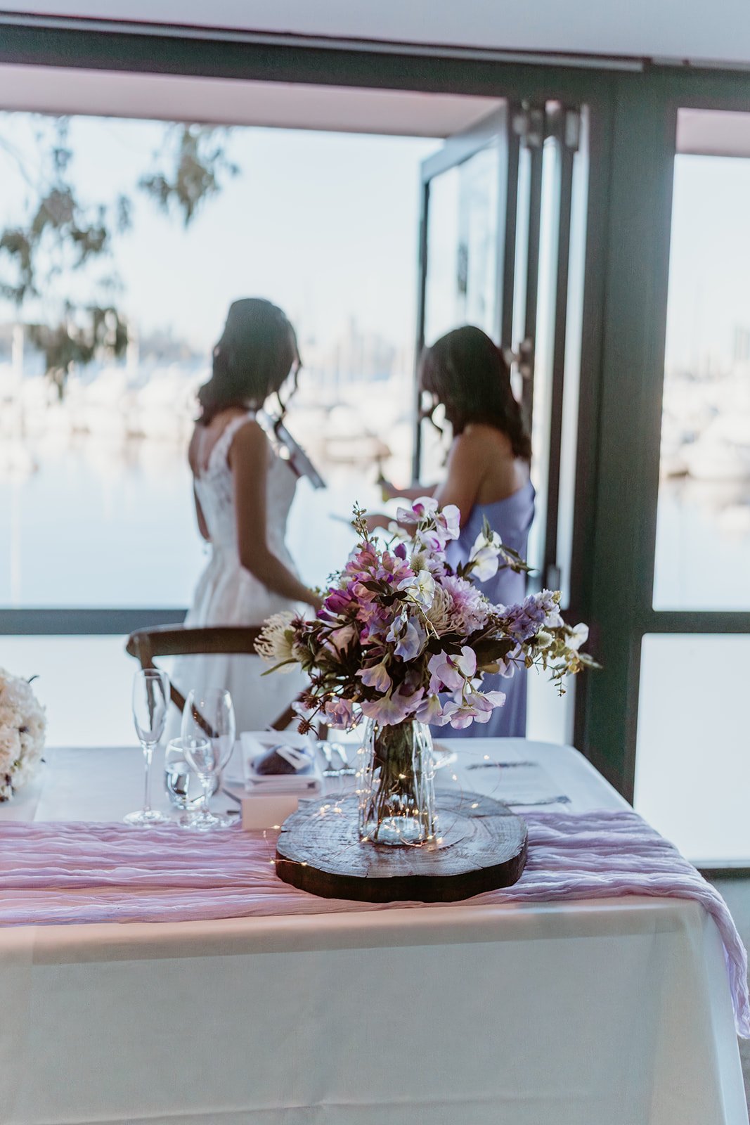 A wedding reception table with a flower arrangement in a glass vase in the foreground, and two women, one in a white dress and the other in purple, standing near a large window overlooking a marina with boats.
