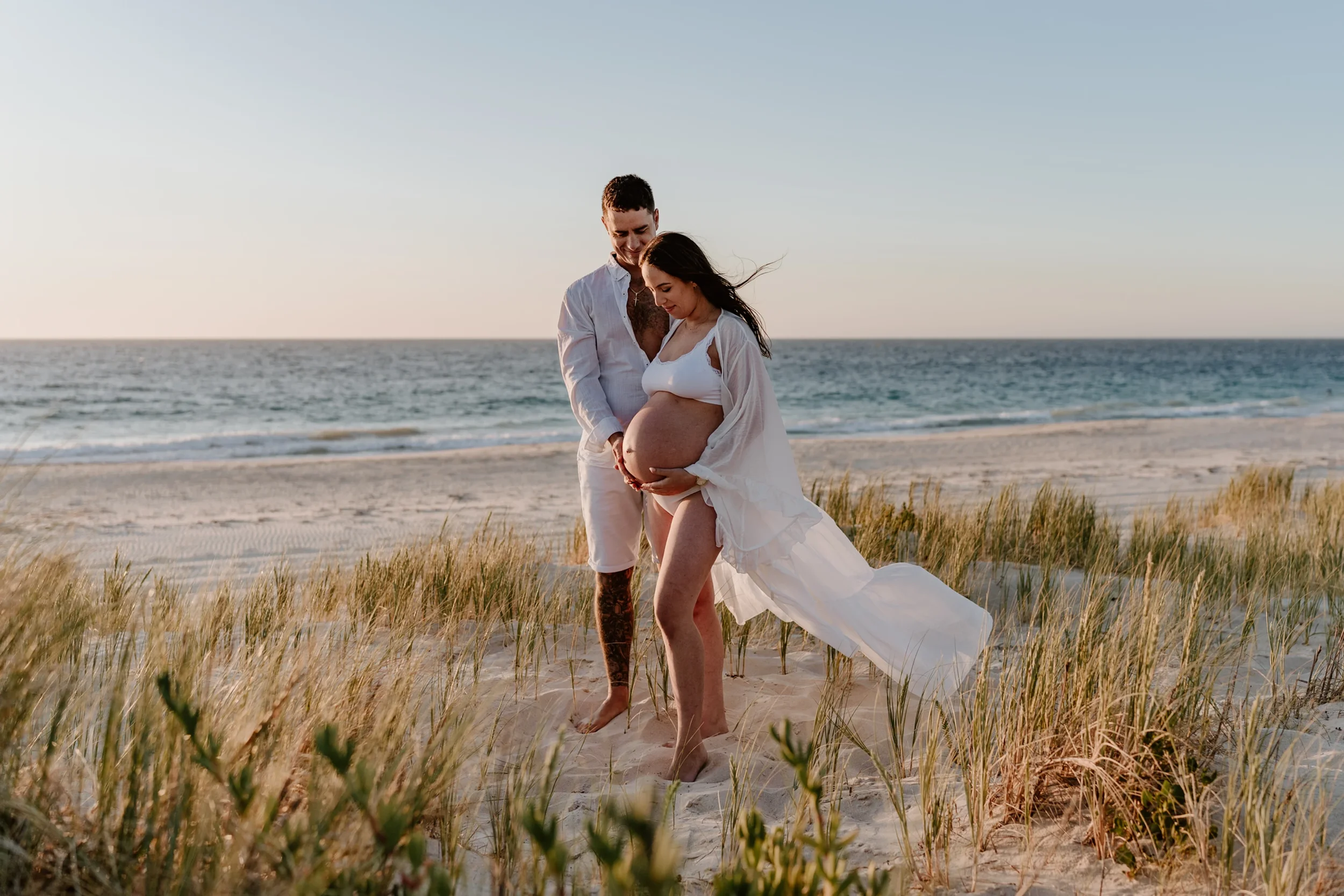 A pregnant woman and a man standing on a beach, holding her belly and touching hands, with the ocean in the background during sunset.