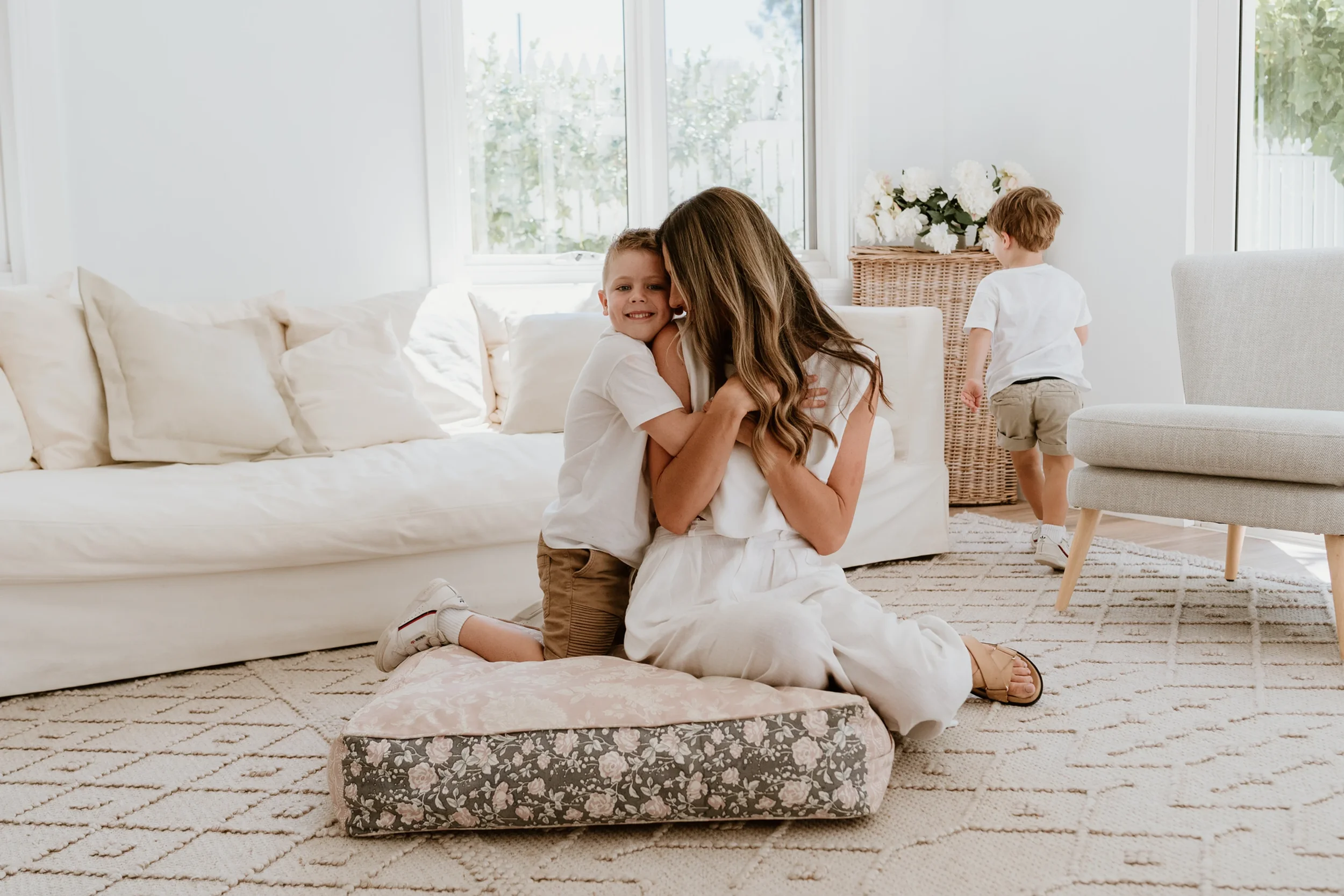 A woman and a boy hugging on a cushion in a bright living room with a white sofa and two children playing in the background.