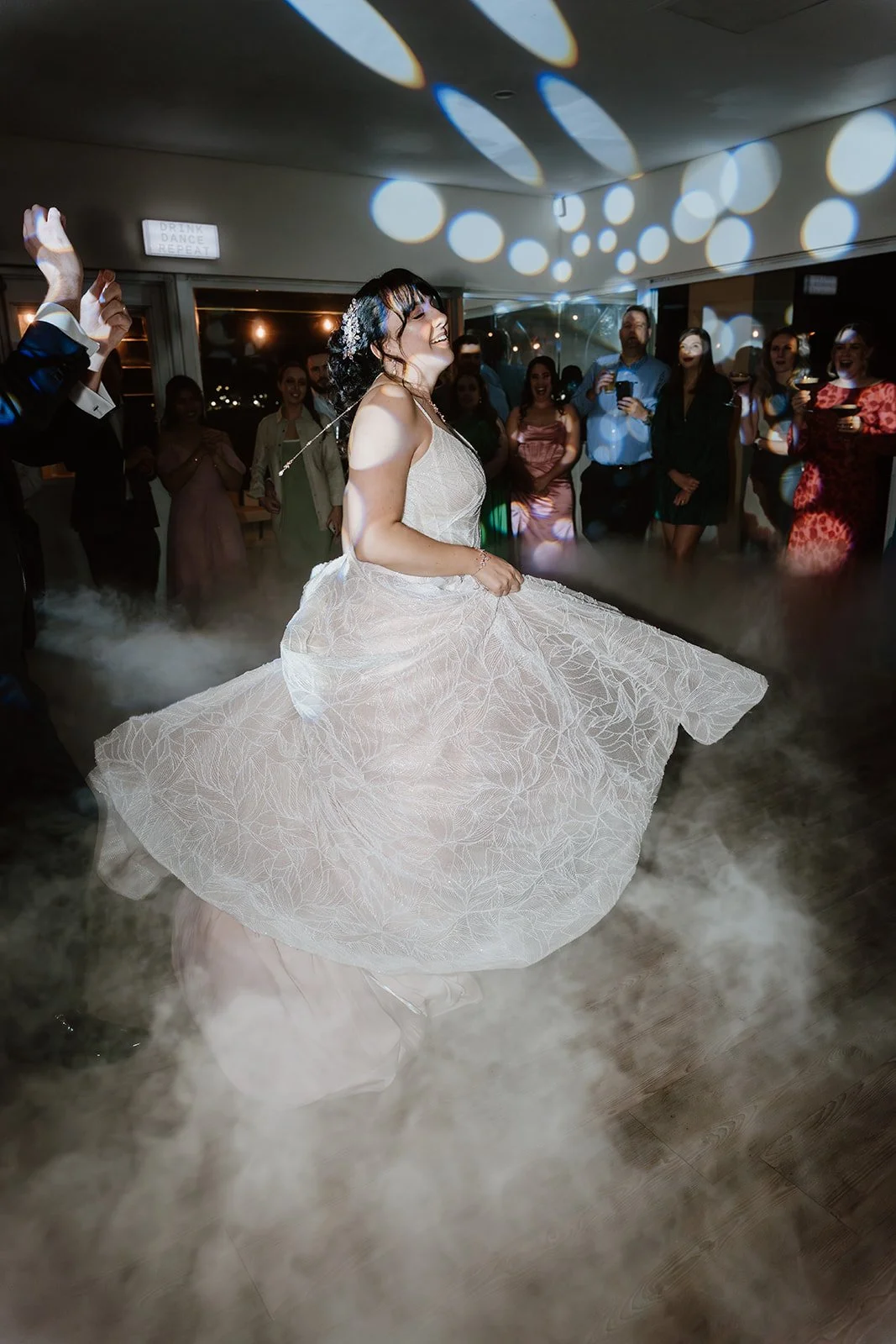 A woman in a white wedding dress dancing at her wedding reception with a big group of guests watching, surrounded by colored lights and fog.