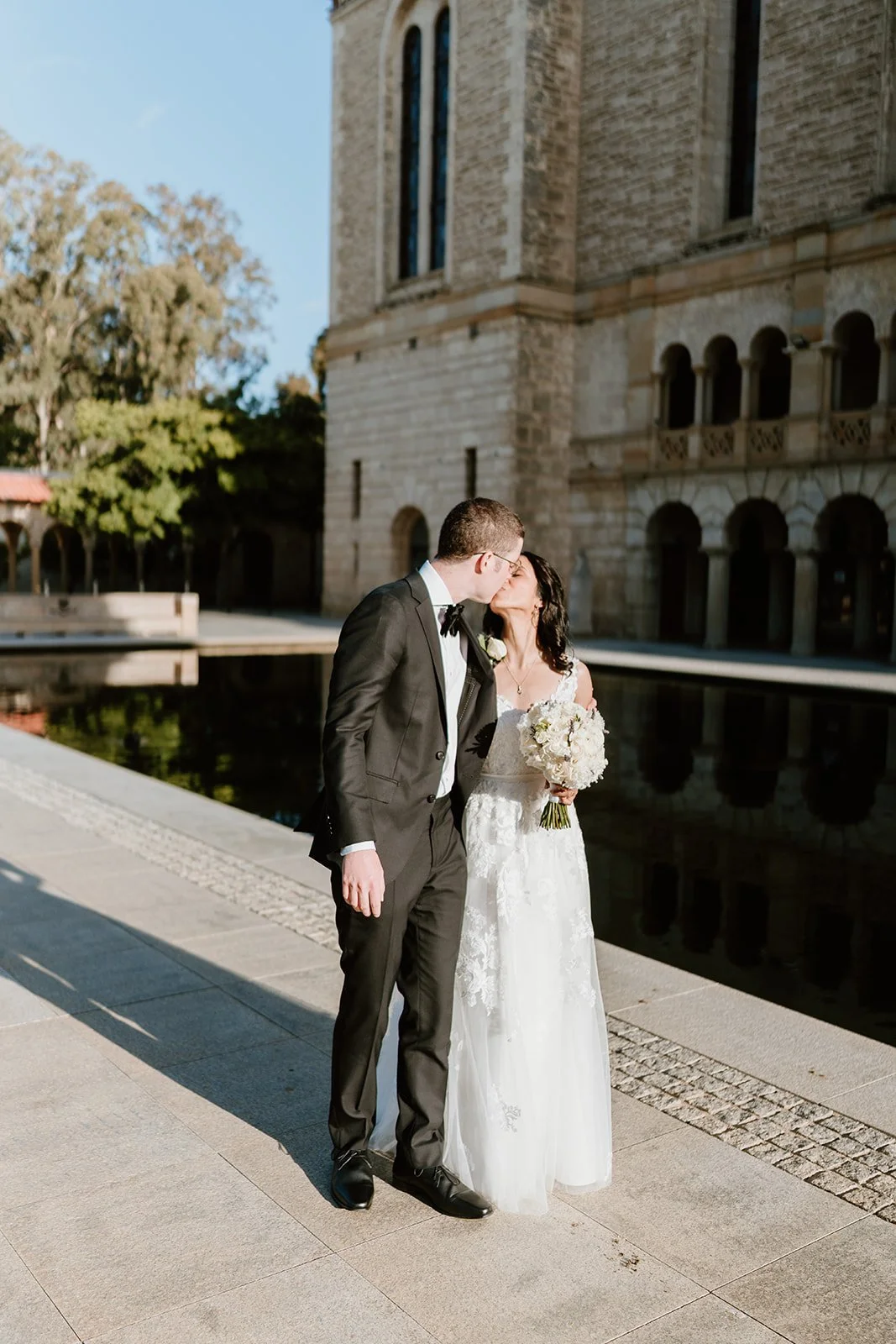 A newlywed couple sharing a kiss outdoors near a historic stone building and reflecting water, with trees and a bright sky in the background.