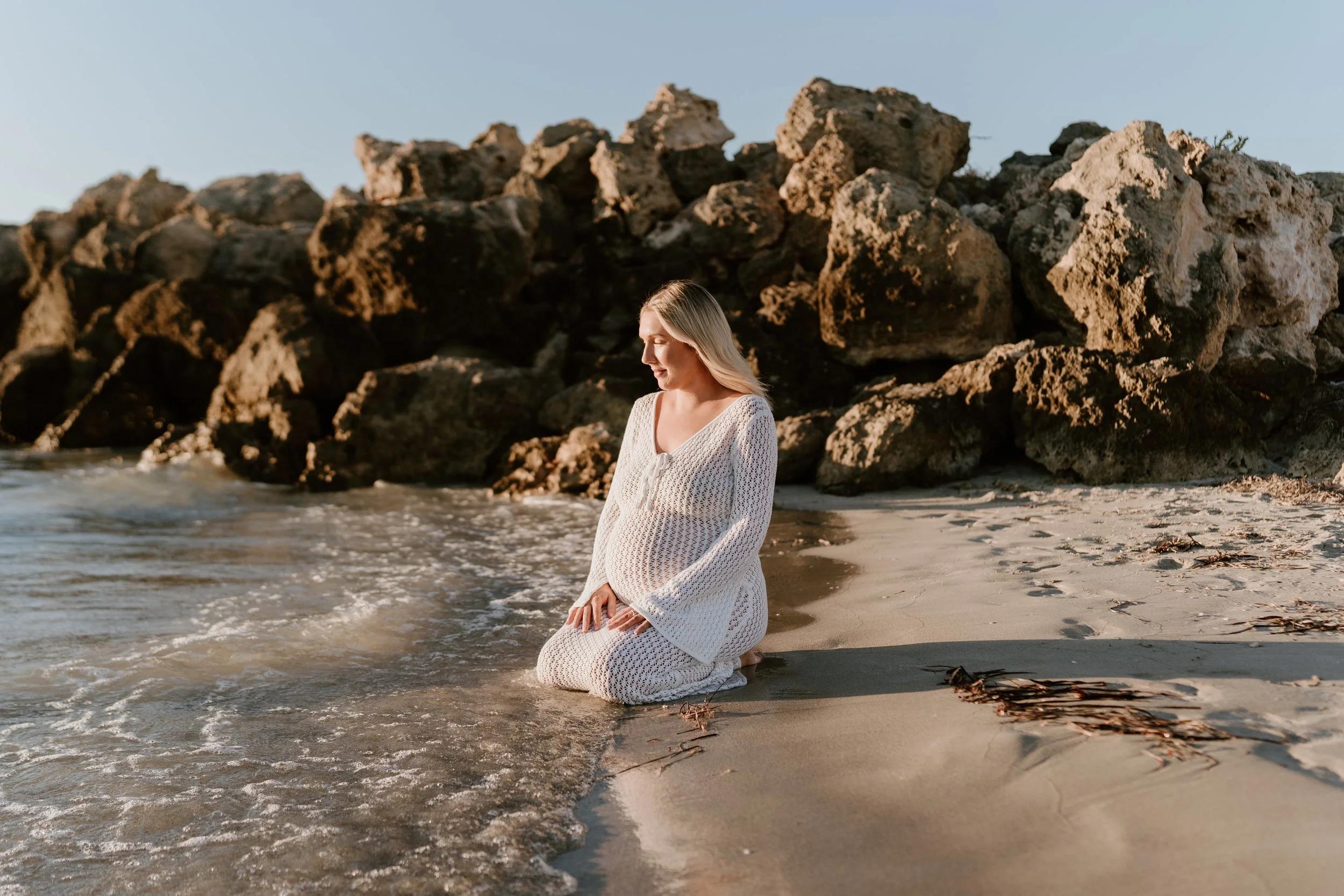 A pregnant woman kneeling on the sandy beach near the water with rocks in the background, illuminated by warm sunlight.