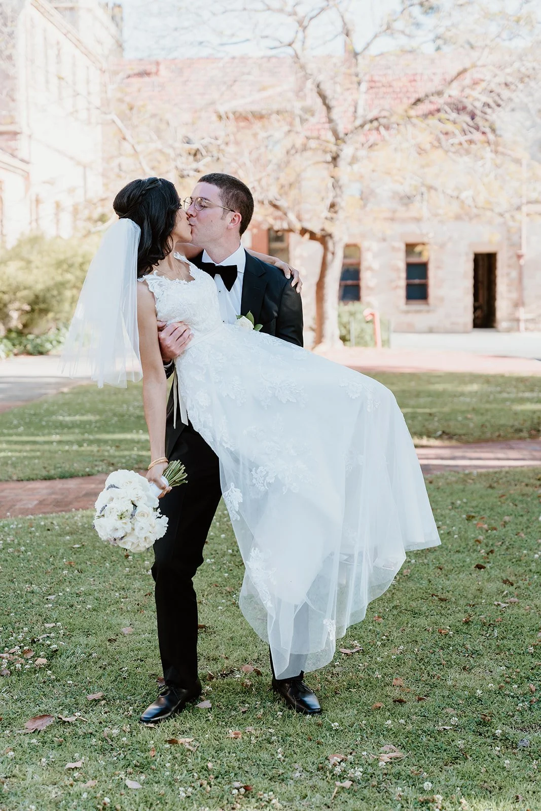 A newlywed couple sharing a kiss outdoors, with the groom lifting the bride. The bride is holding a bouquet of white flowers, and they are dressed in wedding attire on a grassy area with trees and a building in the background.