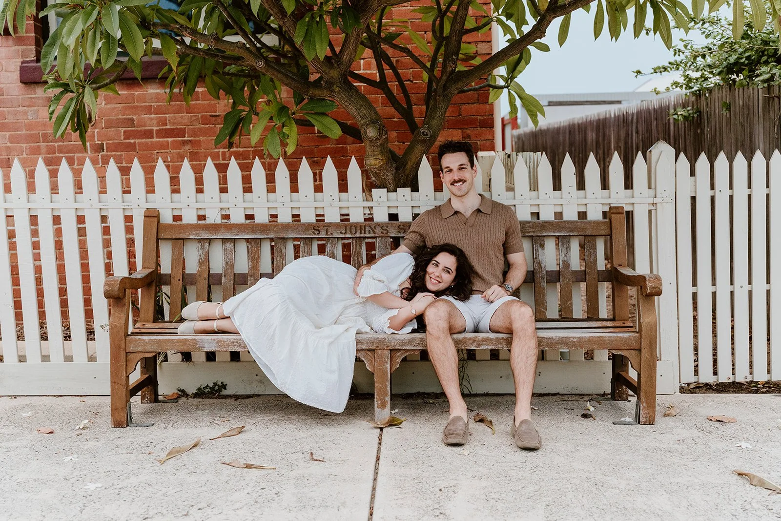 A couple sitting on a wooden bench under a tree in front of a white picket fence. The woman is lying down, resting her head on the man's lap, and smiling, while the man is sitting upright and smiling. There are some fallen leaves on the ground.