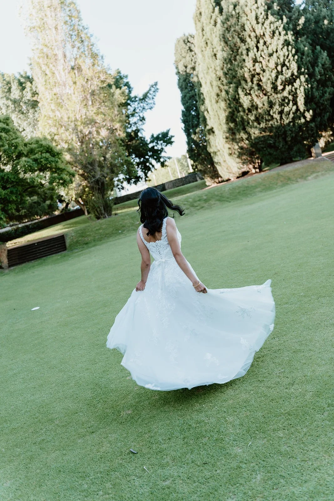 A woman in a white wedding dress walking outdoors on a grassy field, with tall trees and green foliage in the background.