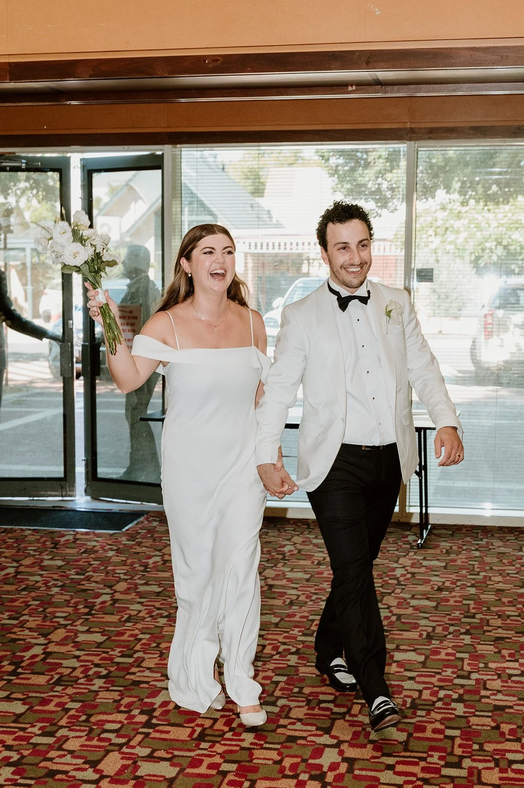 A newlywed couple, the bride in a white off-shoulder dress holding a bouquet, and the groom in a white tuxedo jacket with a black bow tie, holding hands and smiling as they walk into a venue with a patterned carpet and large glass doors.