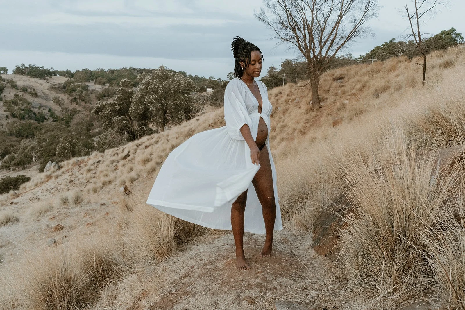 A pregnant woman in a white dress walking barefoot on a dirt path in a grassy, hilly landscape.