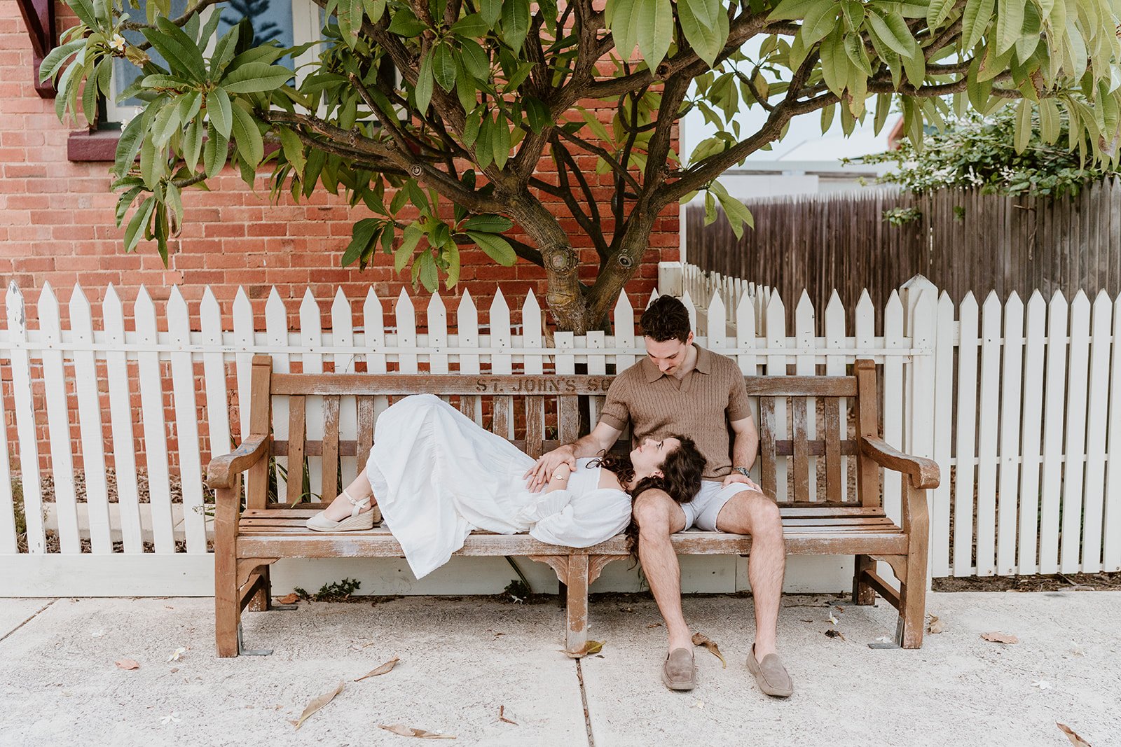 A couple sitting on a park bench beneath a large green tree, with a white picket fence and brick building in the background.