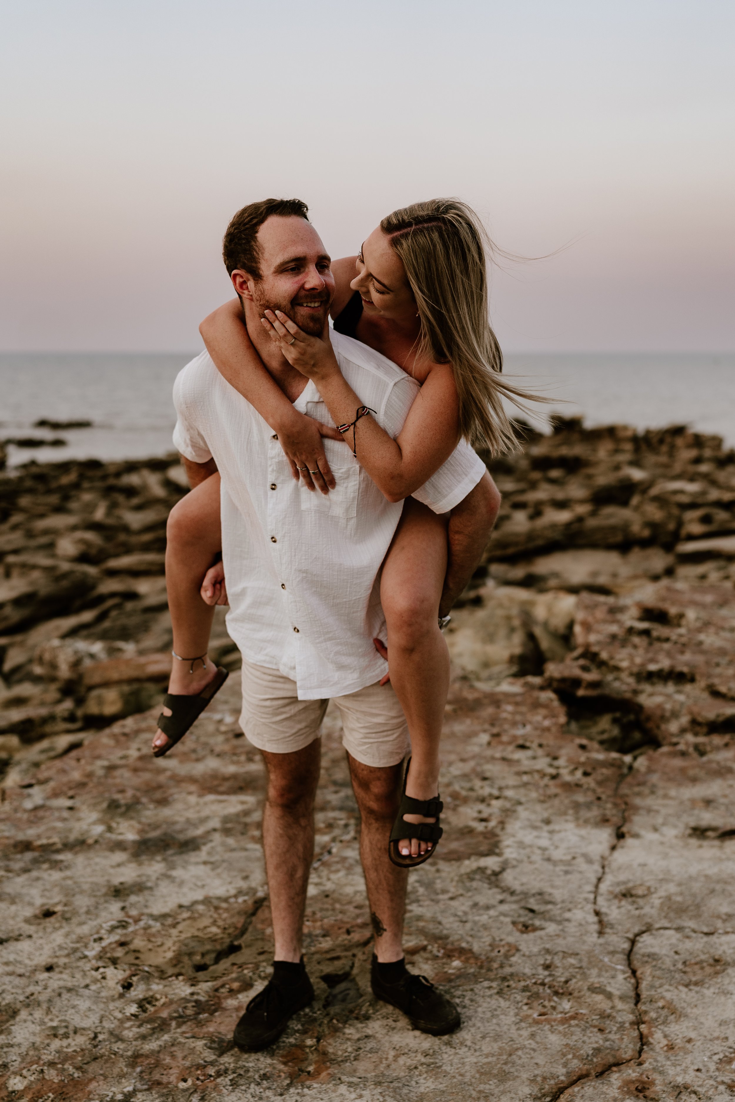 A man giving a piggyback ride to a woman on rocks near the ocean at sunset, both smiling and looking at each other.
