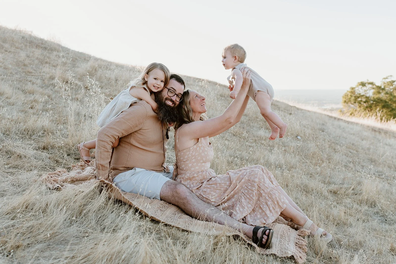 Family of four sitting on a blanket in a grassy field, with the mother holding a toddler in the air and a young girl leaning on the father's back, all smiling.