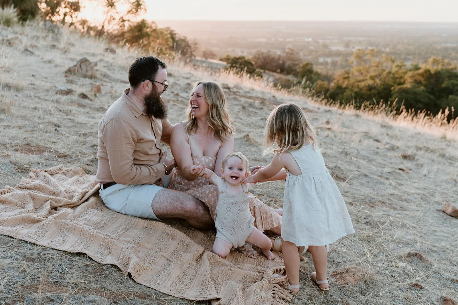 Family of four enjoying time together on a blanket outdoors during sunset, laughing and holding hands, with a scenic landscape background.