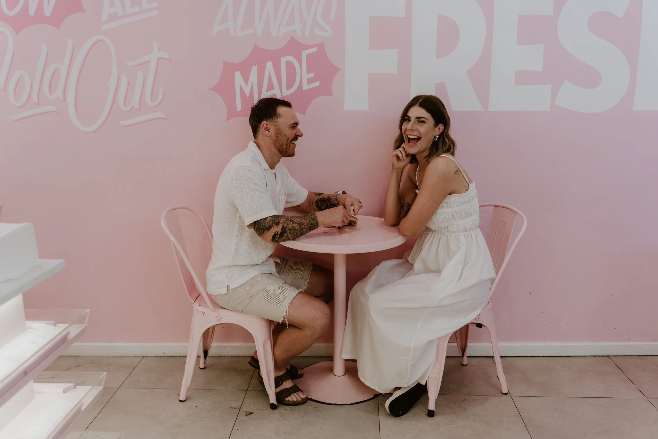 A man and woman sitting at a pink table in a pink-themed space, laughing and enjoying each other's company.
